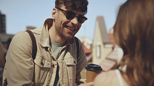 happy man in sunglasses holding paper cup and looking at blurred woman