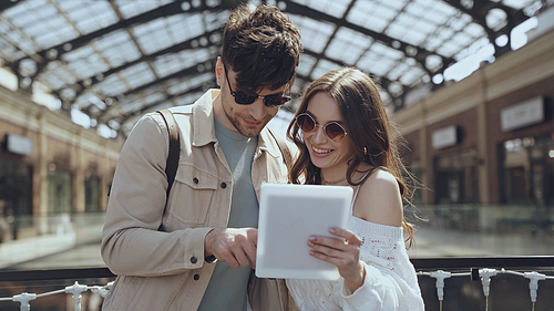 couple in sunglasses smiling while using digital tablet in shopping mall