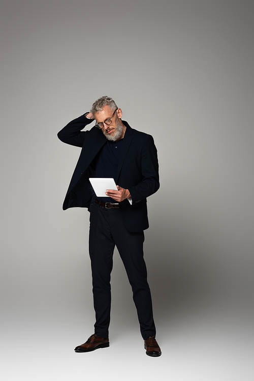 full length of thoughtful middle aged businessman with grey hair looking at digital tablet on grey