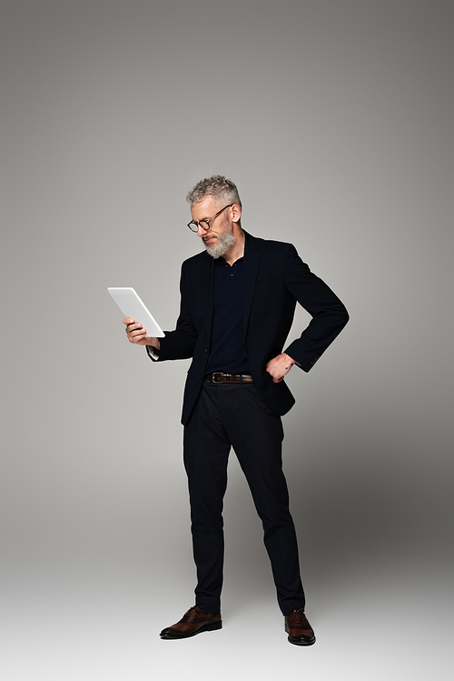 full length of man with grey hair looking at digital tablet and posing on grey