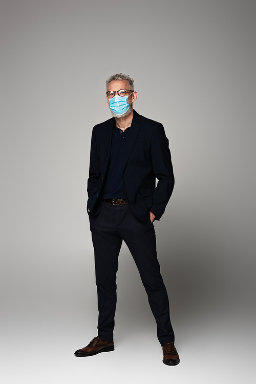 full length of mature man with grey hair in glasses and medical mask posing on grey