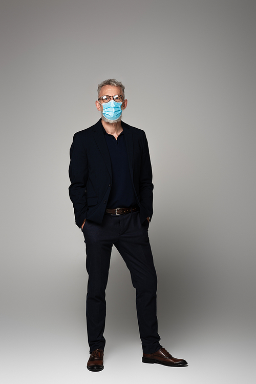 full length of mature man with grey hair in glasses and medical mask posing with hands in pockets on grey