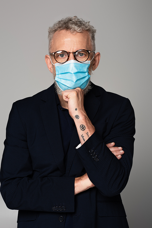 tattooed and mature man with grey hair in glasses and medical mask isolated on grey