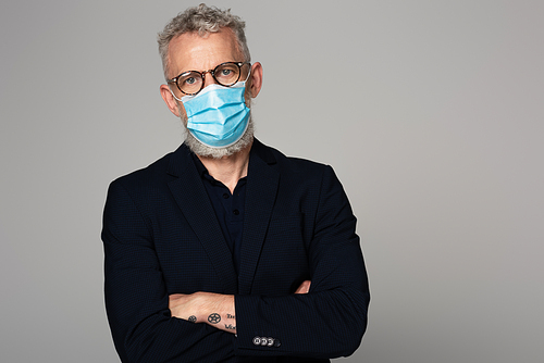 mature man with grey hair in glasses and medical mask standing with crossed arms isolated on grey
