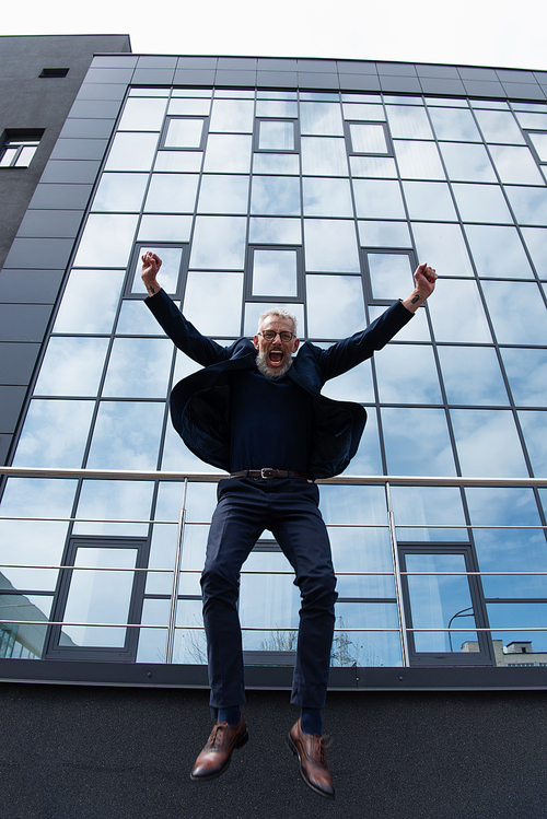 full length of mature man with grey hair in glasses jumping near modern building on grey