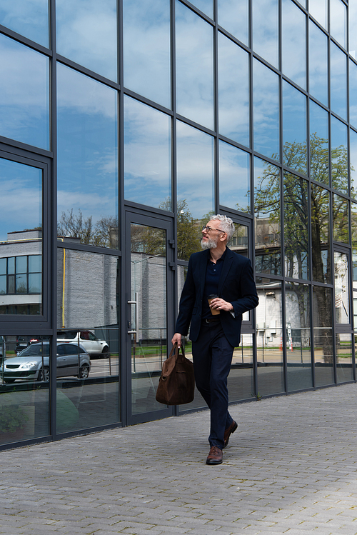 full length of mature businessman with grey hair holding leather bag and paper cup while walking near modern building