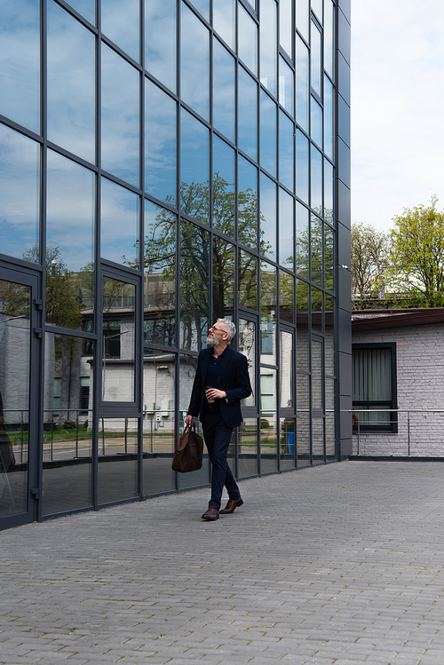 full length of businessman with grey hair holding leather bag and paper cup while walking near modern building