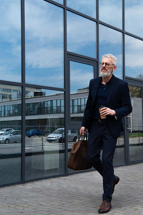 full length of mature man with grey hair holding leather bag and paper cup while walking near modern building