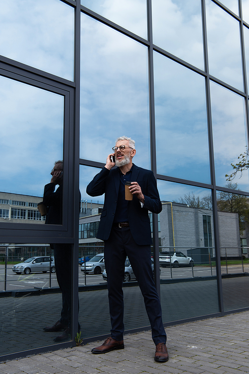 full length of mature man with grey hair holding paper cup while talking on smartphone near modern building