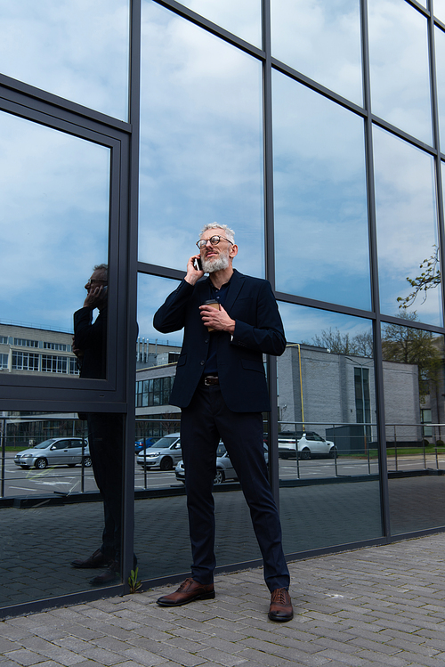 full length of businessman with grey hair holding paper cup while talking on smartphone near modern building