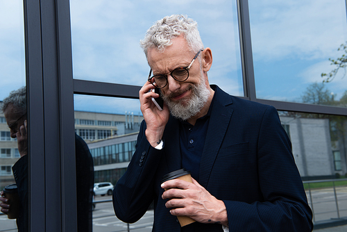 confused businessman with grey hair holding paper cup while talking on smartphone near modern building