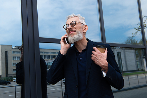 businessman with grey hair holding paper cup while talking on smartphone near modern building