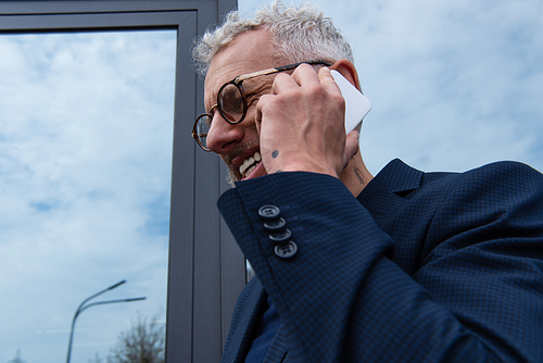 happy businessman with grey hair talking on smartphone near modern building