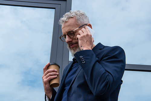 man with grey hair in glasses holding paper cup while talking on smartphone near modern building