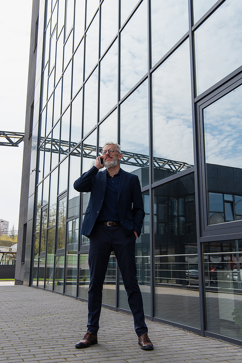 full length of middle aged businessman in glasses talking on smartphone near building