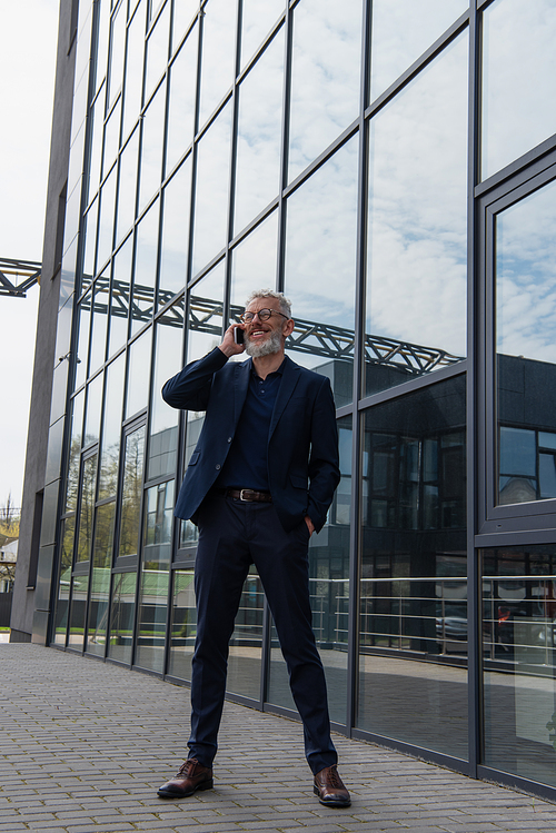 full length of happy middle aged businessman in glasses talking on smartphone near building