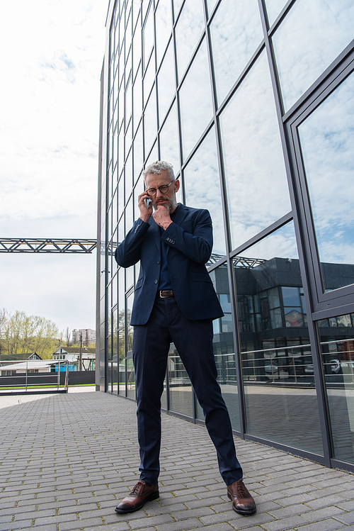full length of pensive middle aged businessman in glasses talking on smartphone near building