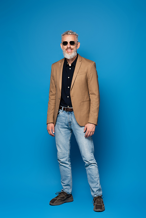 full length of happy middle aged man in sunglasses posing on blue