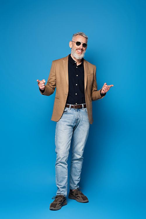 full length of happy middle aged man in sunglasses pointing with fingers on blue