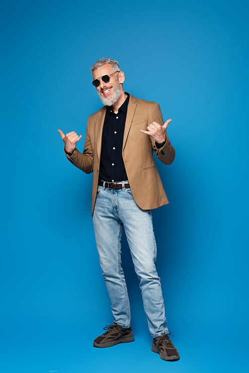 full length of cheerful middle aged man in sunglasses showing call me gesture on blue
