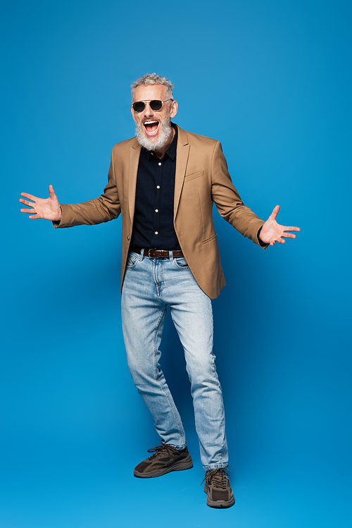 full length of excited middle aged man in sunglasses gesturing on blue