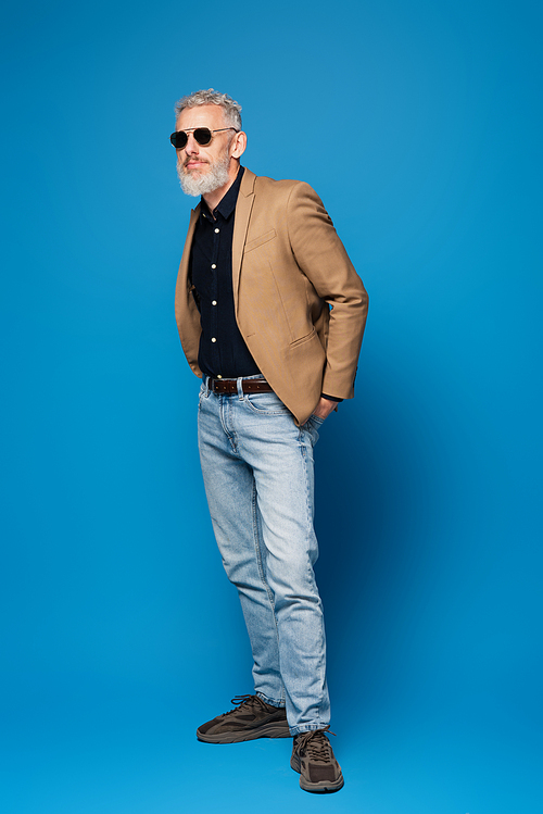 full length of stylish middle aged man in sunglasses posing on blue