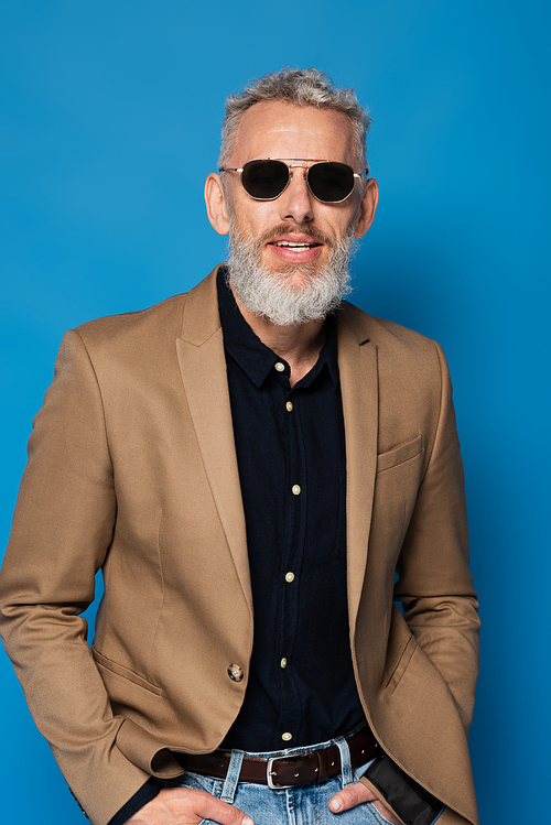 stylish middle aged man in sunglasses standing with hands in pockets isolated on blue
