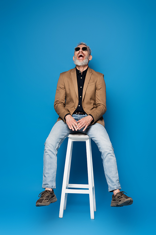 full length of middle aged man in sunglasses laughing on white chair on blue