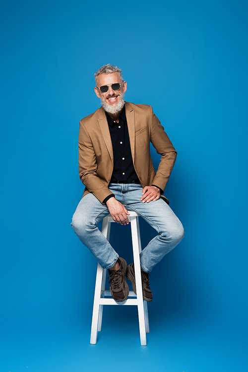full length of cheerful middle aged man in sunglasses posing on white chair on blue
