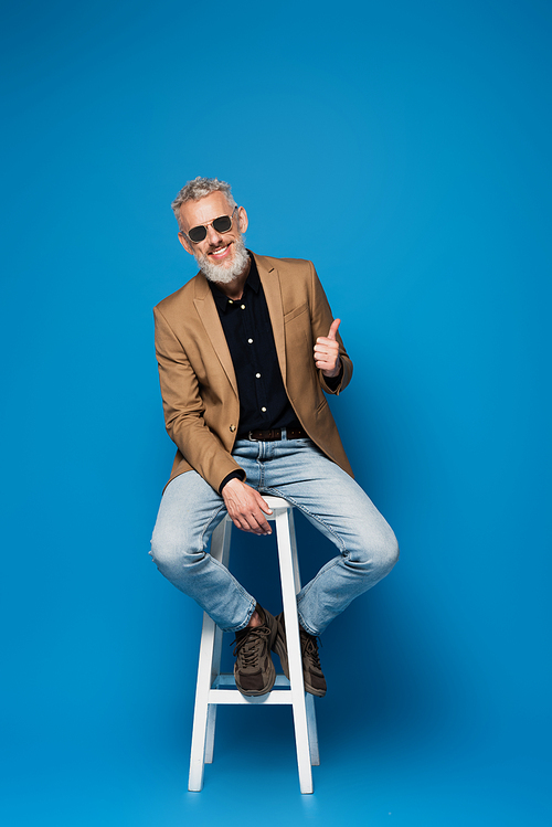 full length of happy middle aged man in sunglasses showing thumb up while sitting on white chair on blue