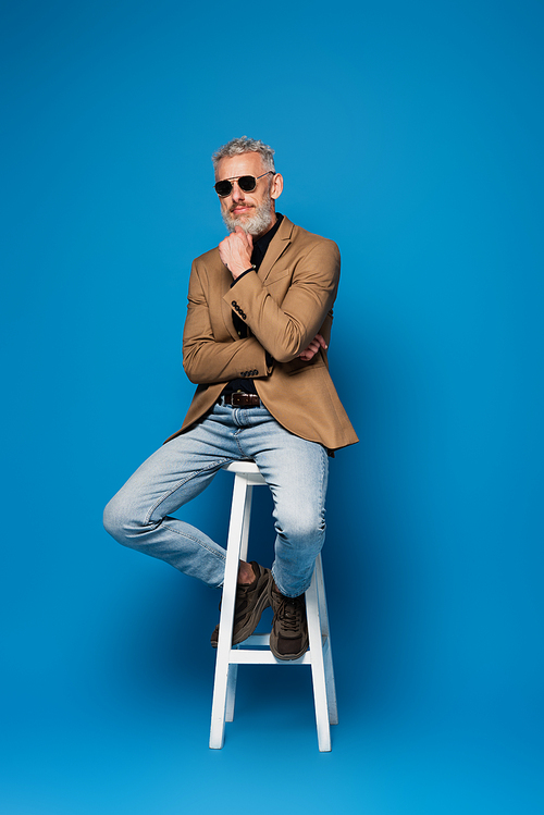 full length of pensive middle aged man in sunglasses posing on white chair on blue