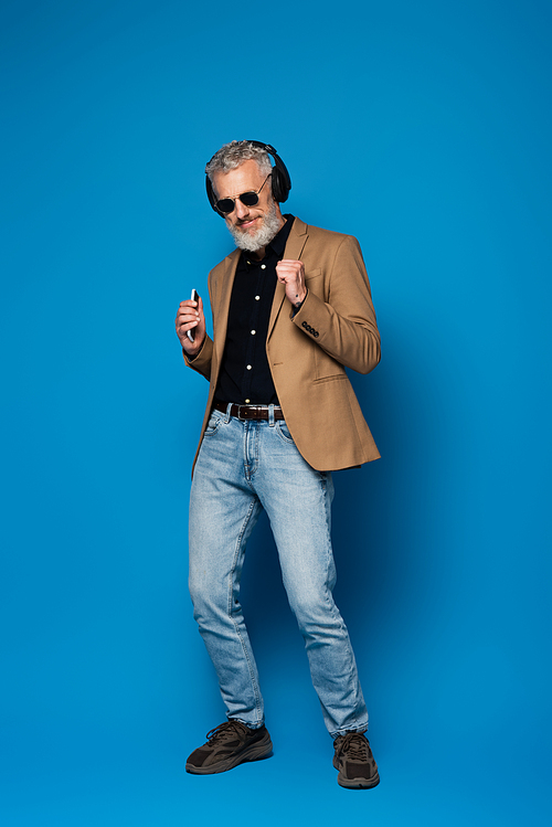 full length of pleased middle aged man in sunglasses holding smartphone and listening music in wireless headphones on blue