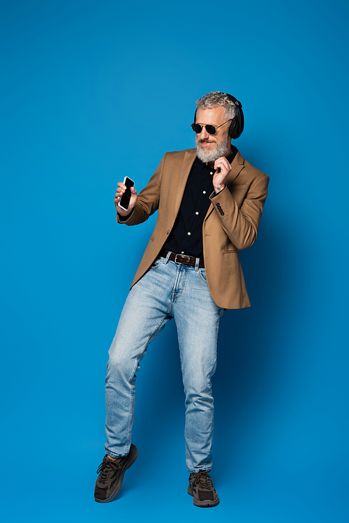 full length of happy middle aged man in sunglasses holding smartphone and listening music in wireless headphones on blue