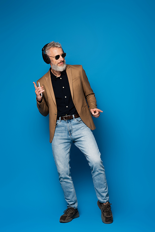 full length of happy middle aged man in sunglasses holding smartphone and listening music in headphones on blue