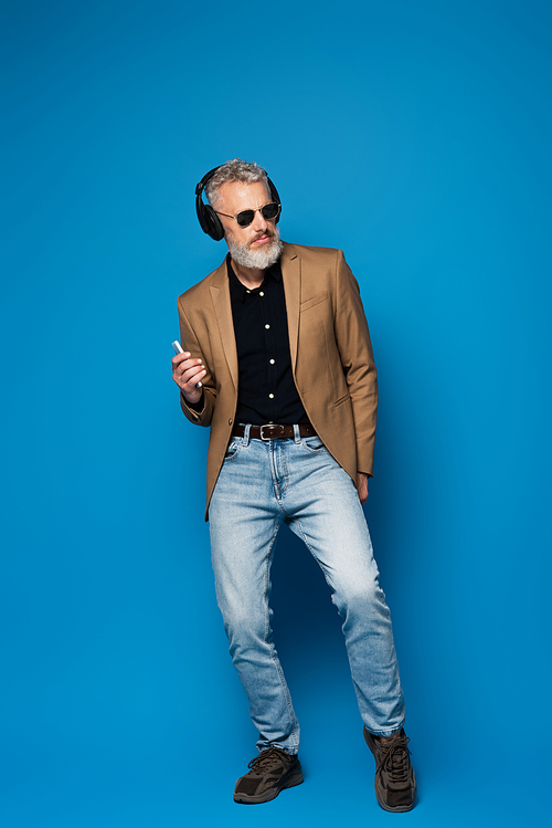full length of middle aged man in sunglasses holding smartphone and listening music in headphones on blue