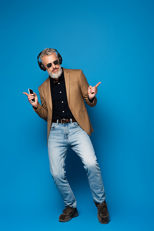 full length of smiling middle aged man in sunglasses holding smartphone while listening music in headphones on blue