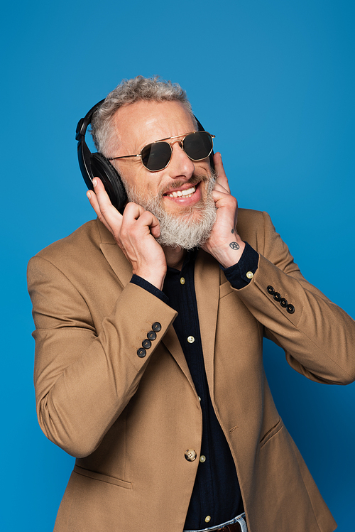 cheerful middle aged man in sunglasses listening music in headphones isolated on blue