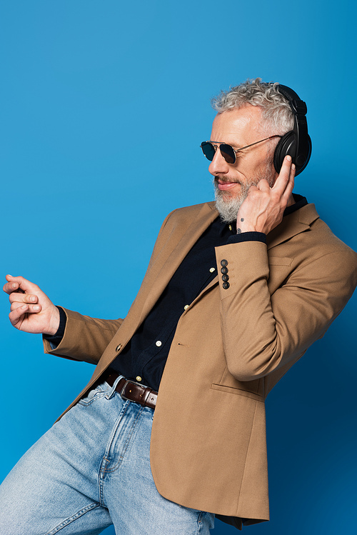 smiling middle aged man in sunglasses listening music in headphones while dancing isolated on blue