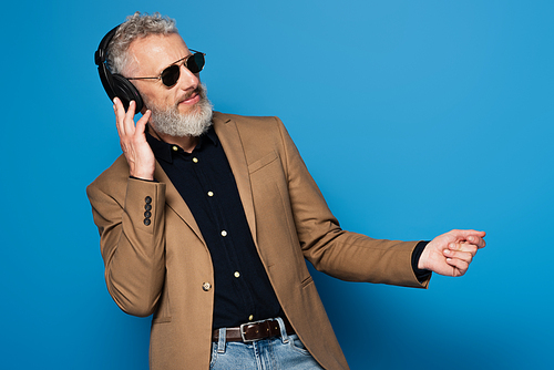 pleased middle aged man in sunglasses listening music in headphones and dancing on blue