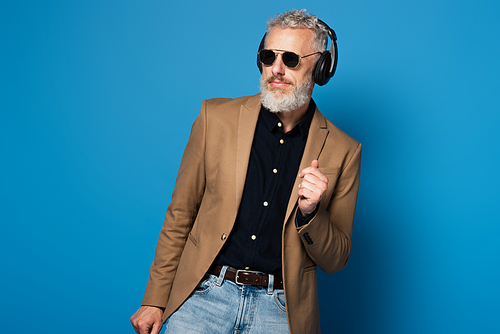 cheerful middle aged man in sunglasses listening music in headphones and dancing on blue