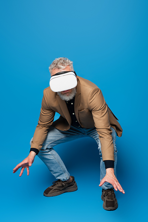full length of middle aged man with grey hair in vr headset squatting and gesturing on blue