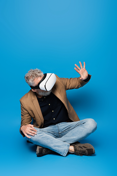 full length of middle aged man in vr headset sitting with crossed legs and gesturing on blue