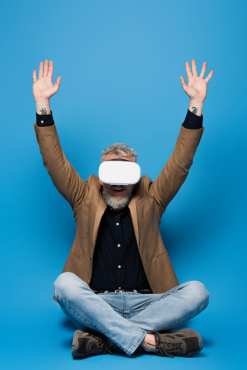full length of excited middle aged man in vr headset sitting with crossed legs and raised arms on blue
