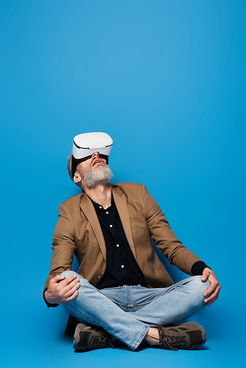 full length of middle aged man in vr headset sitting with crossed legs and looking up on blue