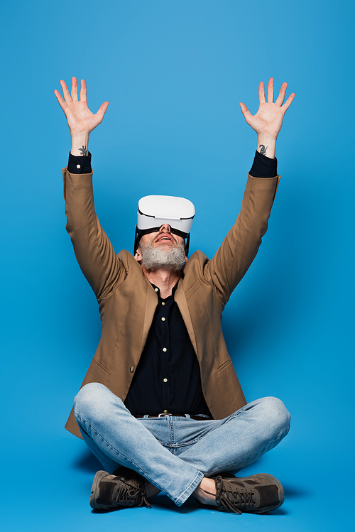 full length of middle aged man in vr headset sitting with crossed legs and raised arms on blue
