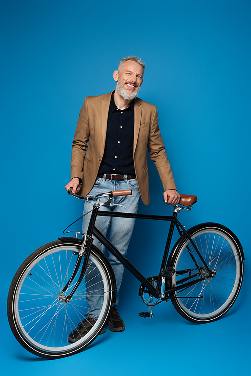full length of pleased middle aged man standing near bicycle on blue
