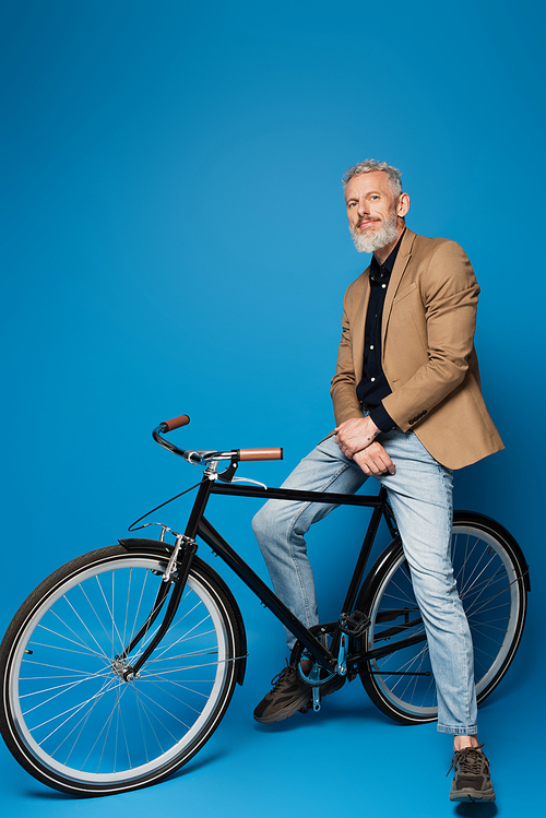 full length of pleased middle aged man sitting on bicycle on blue