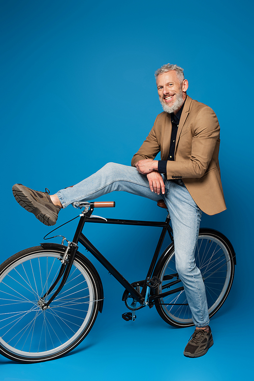 full length of cheerful middle aged man sitting on bicycle on blue