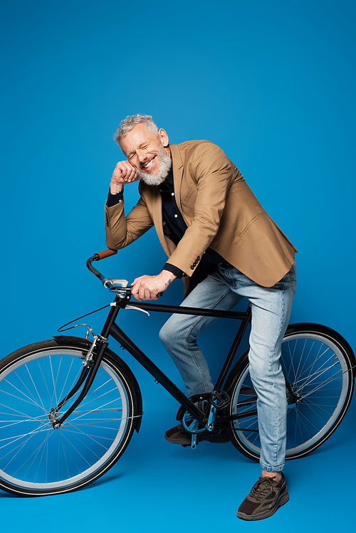 full length of pleased middle aged man sitting on bicycle and smiling on blue
