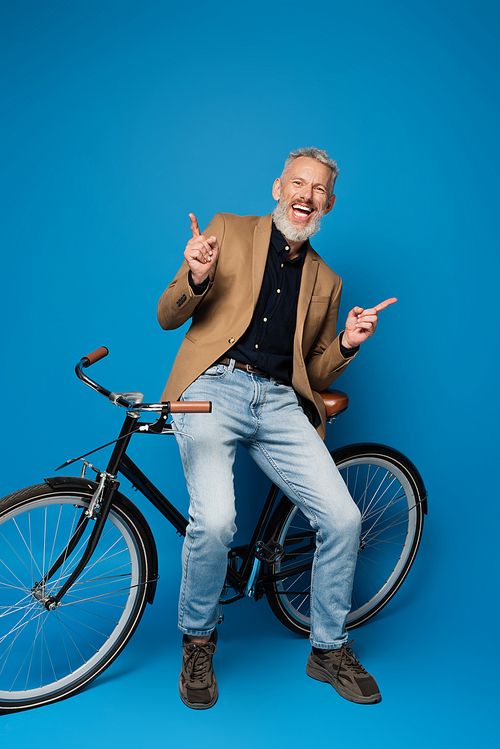 full length of joyful middle aged man sitting on bicycle and pointing with fingers on blue